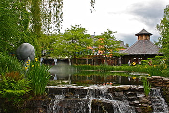 Scenic garden with a pond, waterfall, and sculpture under a cloudy sky. HD wallpaper
