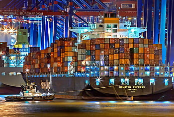 A brightly lit cargo ship at Hamburg harbor with stacked containers and a tugboat. HD wallpaper