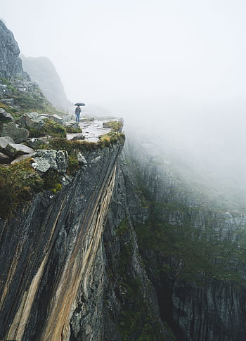 A person with an umbrella stands on a foggy mountain cliff, highlighting travel adventures. HD phone wallpaper