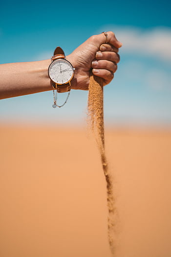 A watch on a hand pouring sand in the Moroccan desert under a clear blue sky. HD phone wallpaper
