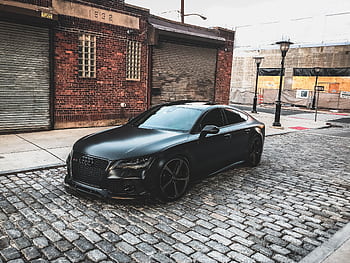 A sleek black car parked on a cobblestone street in an urban setting, featuring brick buildings. HD wallpaper