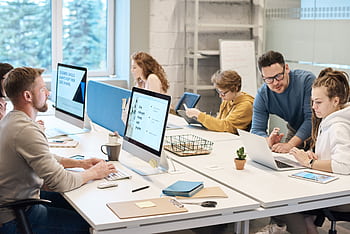 Team collaboration in a modern office setting with computers and diverse employees working together. HD wallpaper