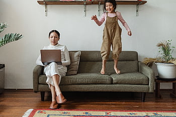 Mother works on laptop at home while child jumps on sofa, showing modern family life. HD wallpaper