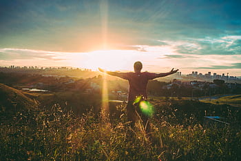 Person standing on grassy hill with open arms embracing a vibrant sunset over a cityscape. HD wallpaper