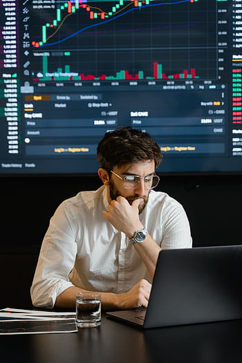 Pensive businessman with a beard analyzing stock market trends on a laptop in an office setting. HD phone wallpaper
