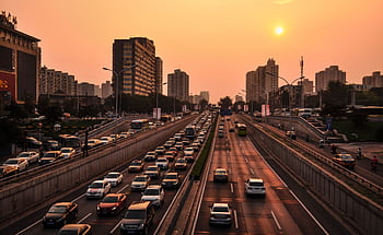 A bustling urban highway with heavy traffic under a vibrant sunset sky against a cityscape backdrop. HD wallpaper
