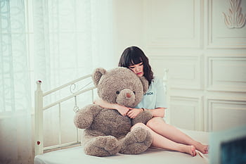 A young woman peacefully hugging a teddy bear while sitting on a bed in a sunlit room. HD wallpaper
