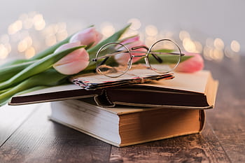 Artistic composition of vintage books, eyeglasses, and pink tulips on a wooden table with bokeh lights. HD wallpaper