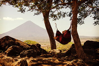 Person relaxing in a hammock between trees with a volcano in the background, capturing tranquility and nature. HD wallpaper