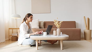 A woman uses a laptop at a coffee table in a cozy living room, embodying a relaxed work-from-home lifestyle. HD wallpaper