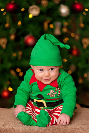 Smiling baby in green elf costume sitting in front of a decorated Christmas tree, capturing holiday joy. HD phone wallpaper