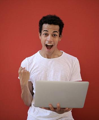 Young man in white shirt with laptop, celebrating success against a vibrant red background. HD phone wallpaper