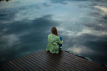 A woman sits on a wooden dock, reflecting by a calm lake under a cloudy sky. HD wallpaper