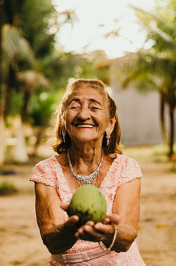 Smiling elderly woman with jewelry showcases happiness while holding a mango outdoors. HD phone wallpaper