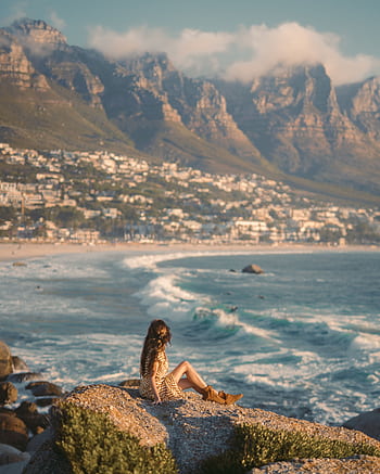 A woman relaxes on rocks overlooking the beautiful coastline of Cape Town, with majestic mountains and a clear blue sky. HD phone wallpaper