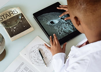 Child exploring moon craters on tablet surrounded by astronomy books and charts. HD wallpaper