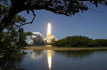 A dramatic rocket launch with smoke and flames, reflected in calm waters and framed by trees. HD wallpaper
