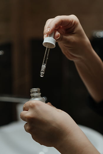 Hands holding a serum dropper applying skincare product in a bathroom setting. HD phone wallpaper