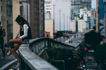 A woman casually sitting on a bridge in a bustling cityscape during the day. HD wallpaper