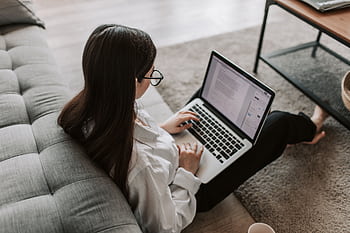 Woman working remotely with a laptop on the floor next to a sofa, enjoying comfortable home office setup. HD wallpaper