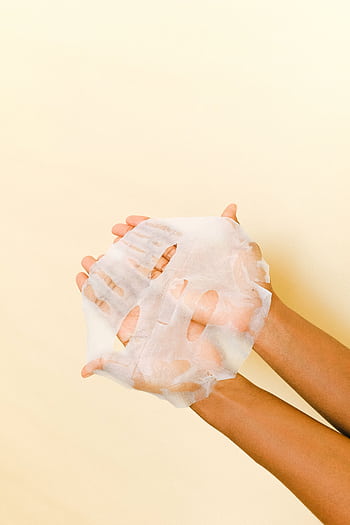 Close-up of hands holding a white sheet mask for skincare on a light background. HD phone wallpaper
