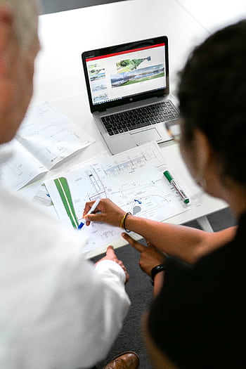 Engineers collaborate on designs with blueprints and laptops in a modern office setting. HD phone wallpaper