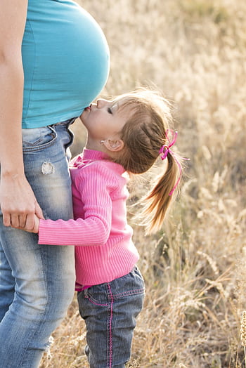 A young girl lovingly kisses her pregnant mother's belly outdoors, symbolizing family affection. HD phone wallpaper