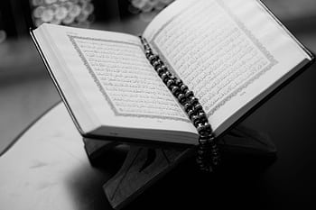 A black and white close-up of an open Quran on a wooden stand with prayer beads, symbolizing Islamic faith and devotion. HD wallpaper