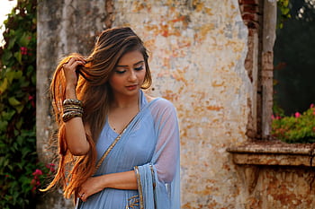 A woman in a blue saree poses gracefully outdoors by a rustic wall. HD wallpaper