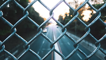 A blurred view of a highway through a chain-link fence at sunset, capturing the essence of urban transit. HD wallpaper