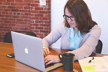 Young woman working on a laptop at a modern office desk with brick wall. HD wallpaper