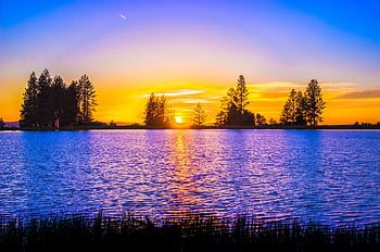 A breathtaking view of a vibrant sunset over a serene lakeside with lush silhouettes of trees. HD wallpaper