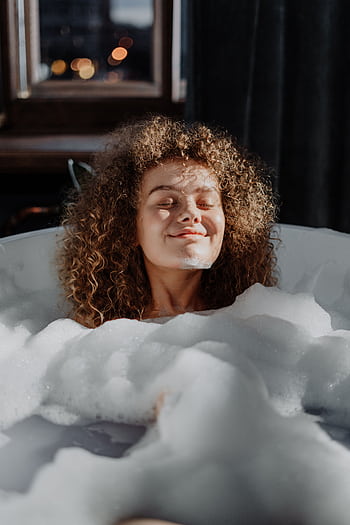 A young woman enjoys a relaxing bubble bath with natural light enhancing her curly hair. HD phone wallpaper