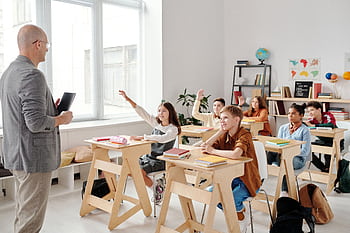 Students engage eagerly in a classroom, raising hands and participating in a lesson with their teacher. HD wallpaper