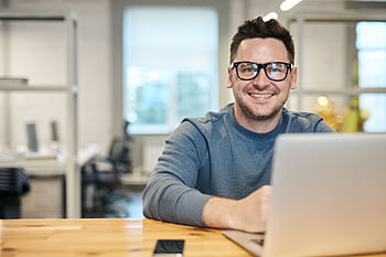 Happy man wearing glasses working remotely on laptop in modern office environment. HD wallpaper
