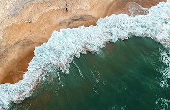 Aerial shot of ocean waves crashing onto a sandy beach with two people in the distance. HD wallpaper