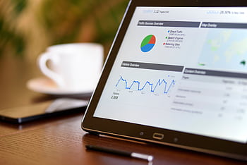 Close-up of a tablet displaying analytics charts on a wooden office desk, alongside a smart and coffee cup. HD wallpaper