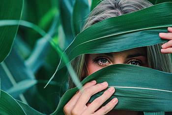 Close-up portrait of a young woman peeking through lush green leaves, in an outdoors setting. HD wallpaper
