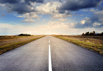 A long straight road stretches through fields under a dramatic cloudy sky. HD wallpaper