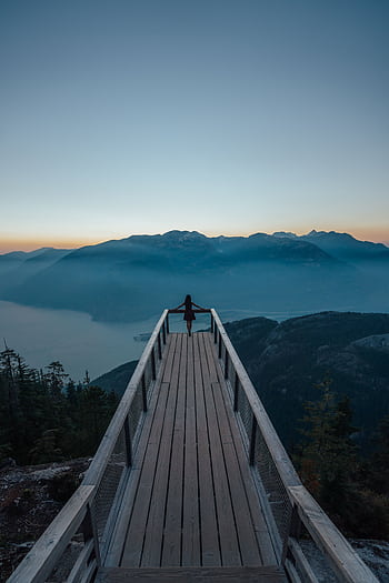 A lone person stands on a wooden boardwalk, gazing into the foggy mountain range, under a clear sky at dawn. HD phone wallpaper