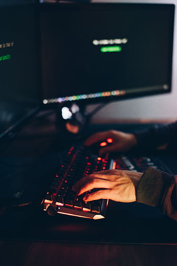 Hands typing on a backlit keyboard with blurred monitors in a dimly lit room. HD phone wallpaper