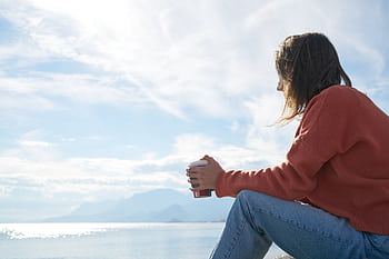 A woman in casual attire enjoys a hot drink while gazing at the sea, epitomizing relaxation. HD wallpaper
