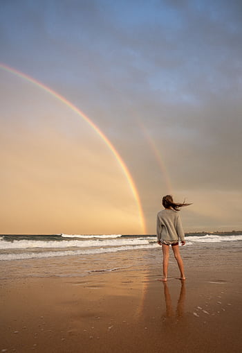 A woman stands barefoot on a beach shore gazing at a vibrant double rainbow under a dramatic sky. HD phone wallpaper