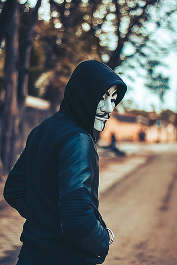 A man in a hoodie wearing a Guy Fawkes mask walks down an urban street, creating an air of mystery and intrigue. HD phone wallpaper