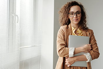 Curly-haired businesswoman wearing glasses holds a laptop in a bright office setting. HD wallpaper