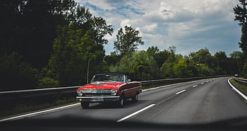 Vintage red convertible cruising along a scenic highway with lush greenery. HD wallpaper
