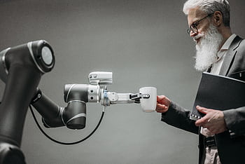 An elderly man receives a cup from a robotic arm in a modern office setting. HD wallpaper