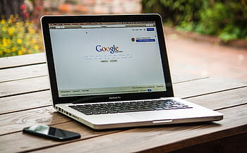 A MacBook Pro displaying Google Search on a wooden table outdoors, next to a smart. HD wallpaper