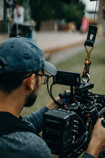 A focused filmmaker operating a cinema camera outdoors during daylight, capturing a creative video shoot. HD phone wallpaper
