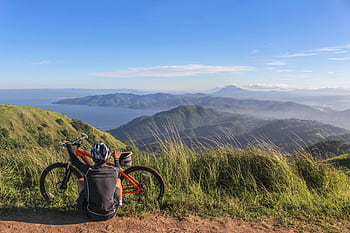 A mountain biker resting by their bike, enjoying a scenic view of lush hills and distant water under a bright blue sky. HD wallpaper
